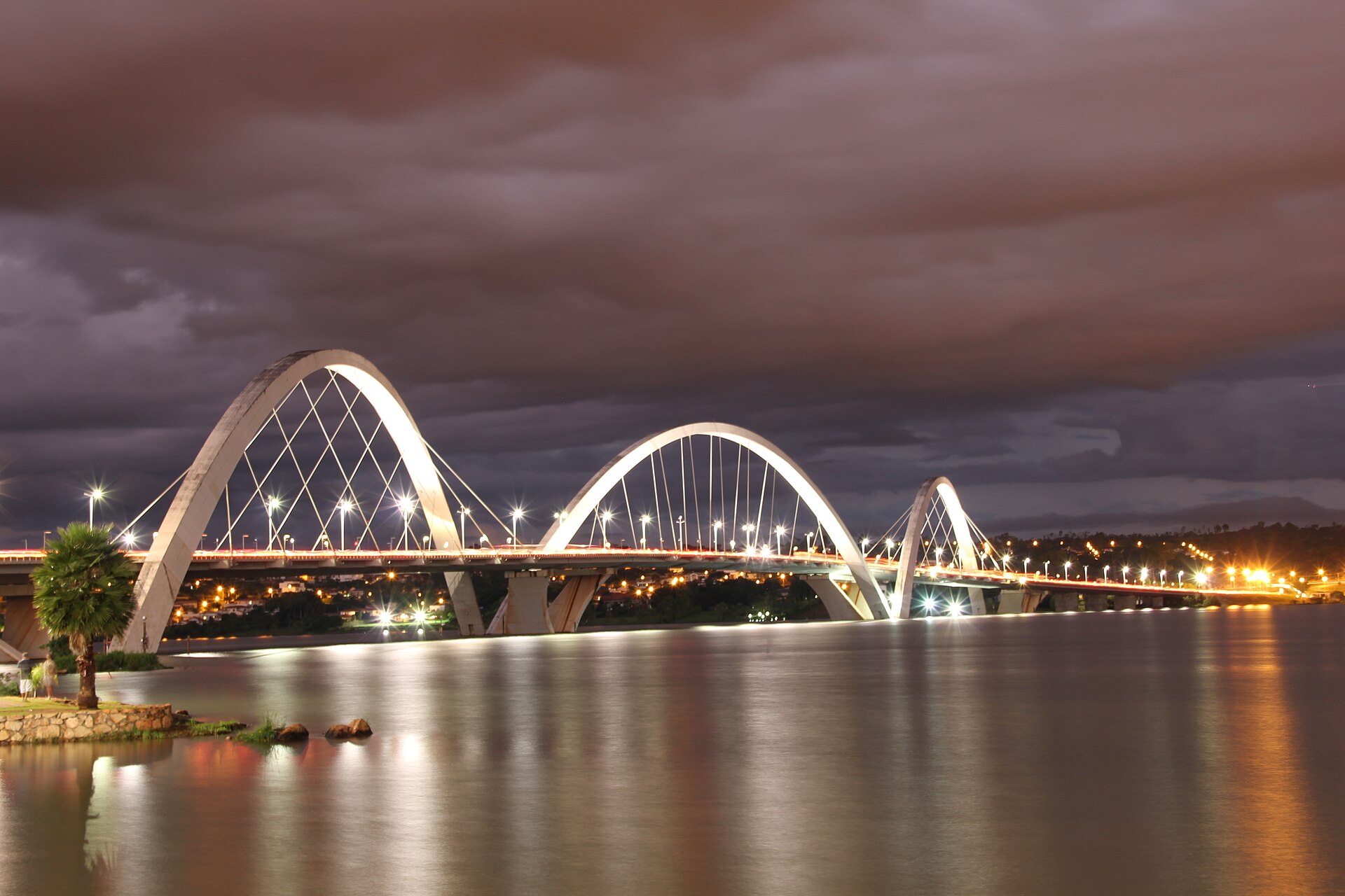 The white arches of the JK Bridge lit at dusk over the still water of Lake Paranoá under a dark cloudy sky