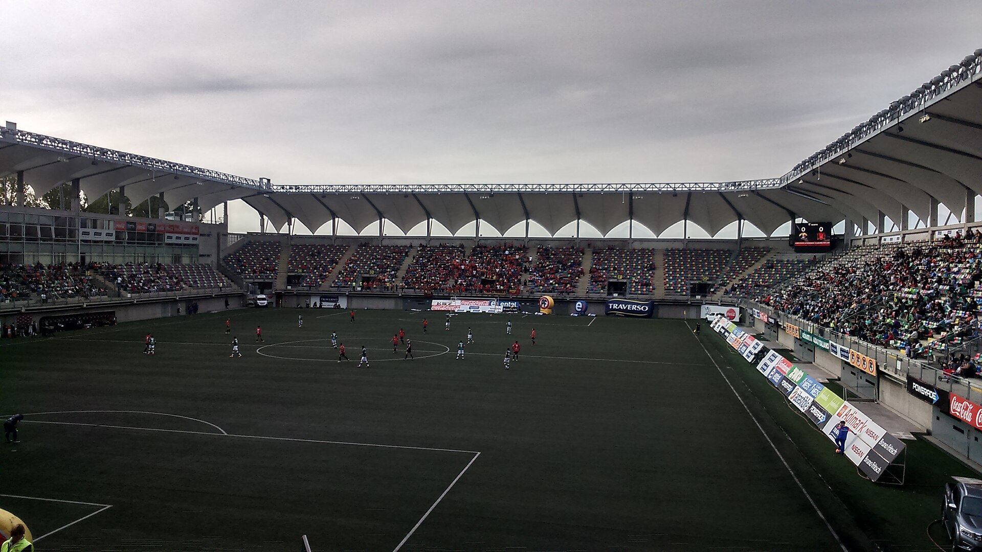 Football match on the pitch of Estadio Bicentenario de La Florida under a grey sky, with white roof canopies over the stands