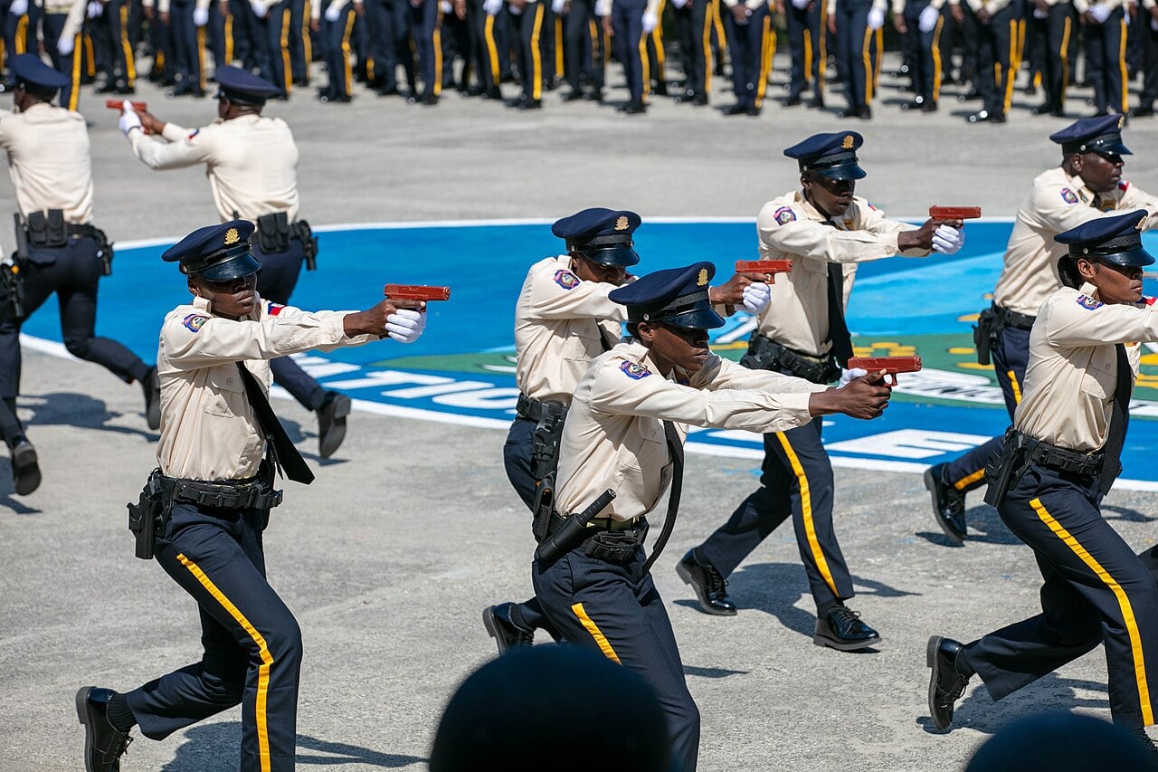 Haitian National Police cadets in cream shirts and navy caps point training pistols during a drill on a parade ground