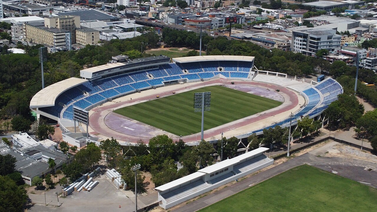 Aerial view of an empty stadium with blue seats, a green pitch and a running track, surrounded by trees and city buildings