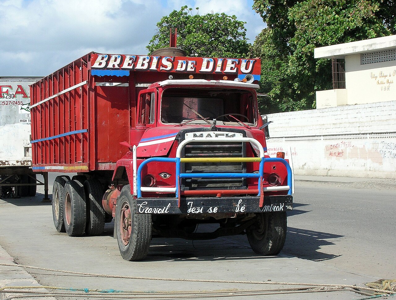 A red Mack cargo truck with a painted wooden body and a slogan on its bumper parked on a street in Cap-Haïtien.