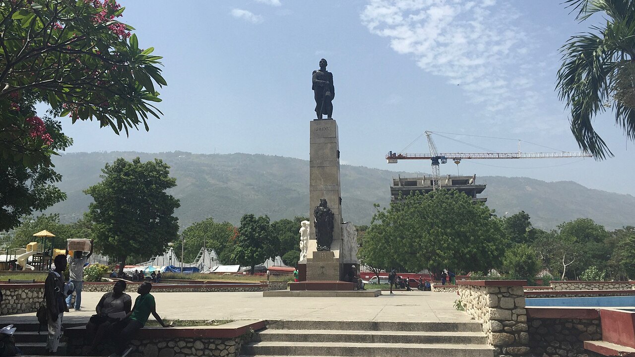 A public square with a statue on a tall plinth in Port-au-Prince, with green hills behind the city