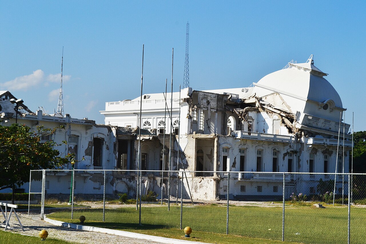 The damaged white dome and walls of the National Palace in Port-au-Prince behind a wire fence, under a blue sky