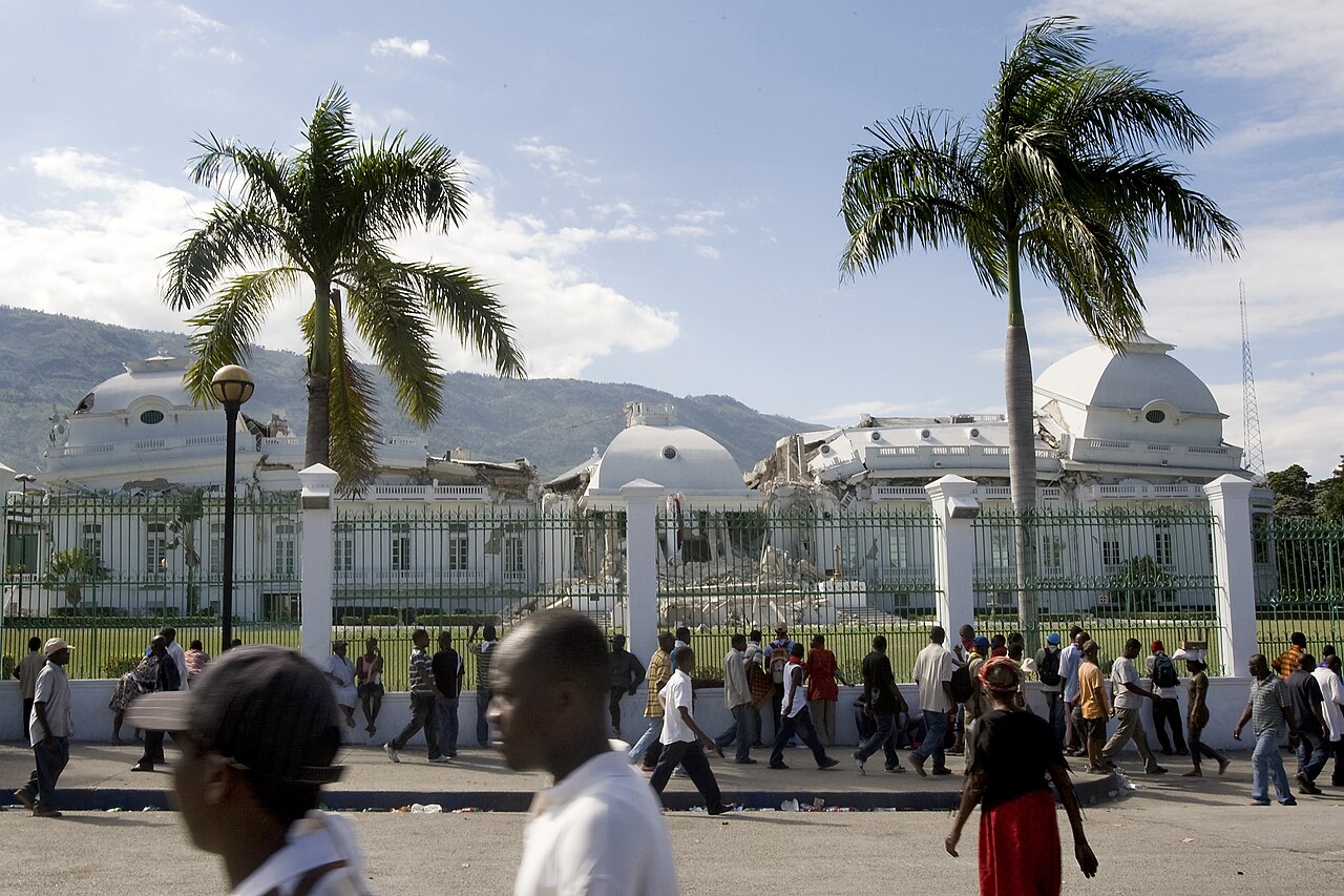 Pedestrians walk past the green iron fence of the damaged National Palace in Port-au-Prince, with two palm trees and white domes behind
