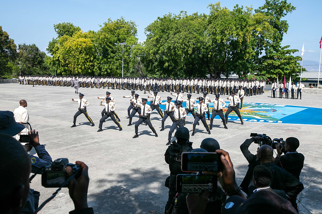 Uniformed Haitian National Police cadets in a drill display on a parade ground while rows of officers stand behind and cameras film
