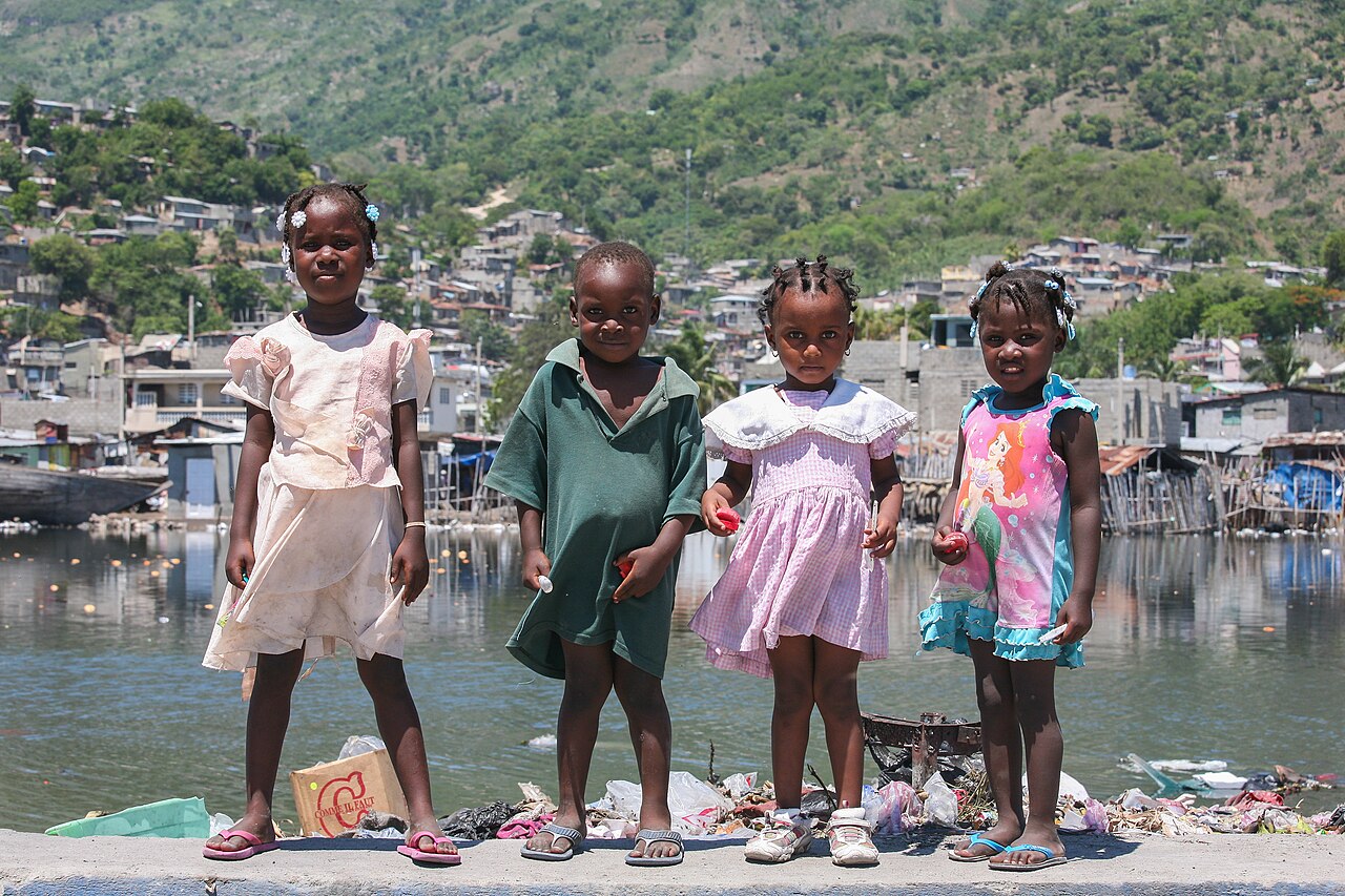 Four young children stand on a harbour wall in Haiti, with houses on green hills behind them