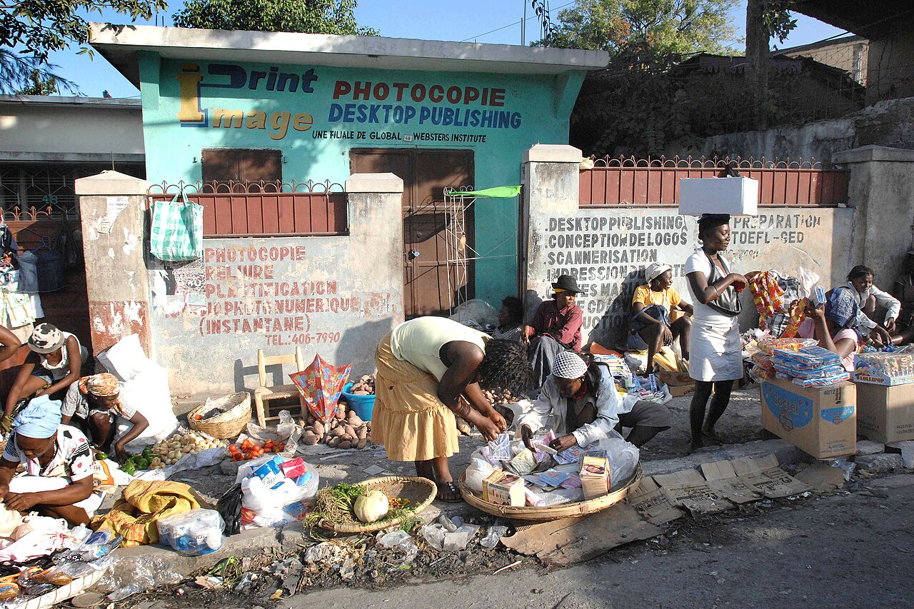 Women sell vegetables and goods on a roadside market in front of a painted shop wall in Port-au-Prince
