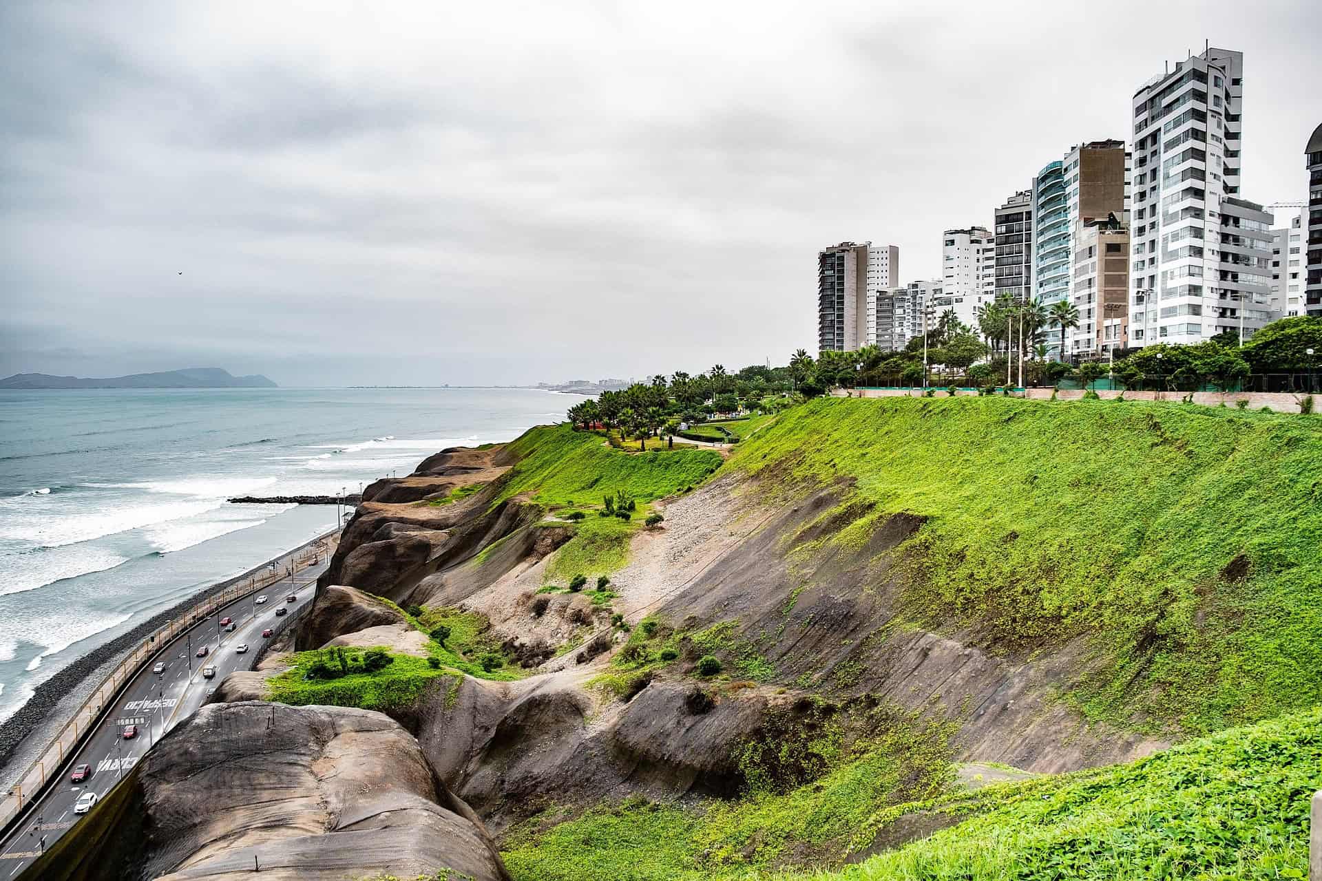 Green cliffs, apartment towers and a coastal highway beside the Pacific Ocean on the Lima coast under a grey sky.