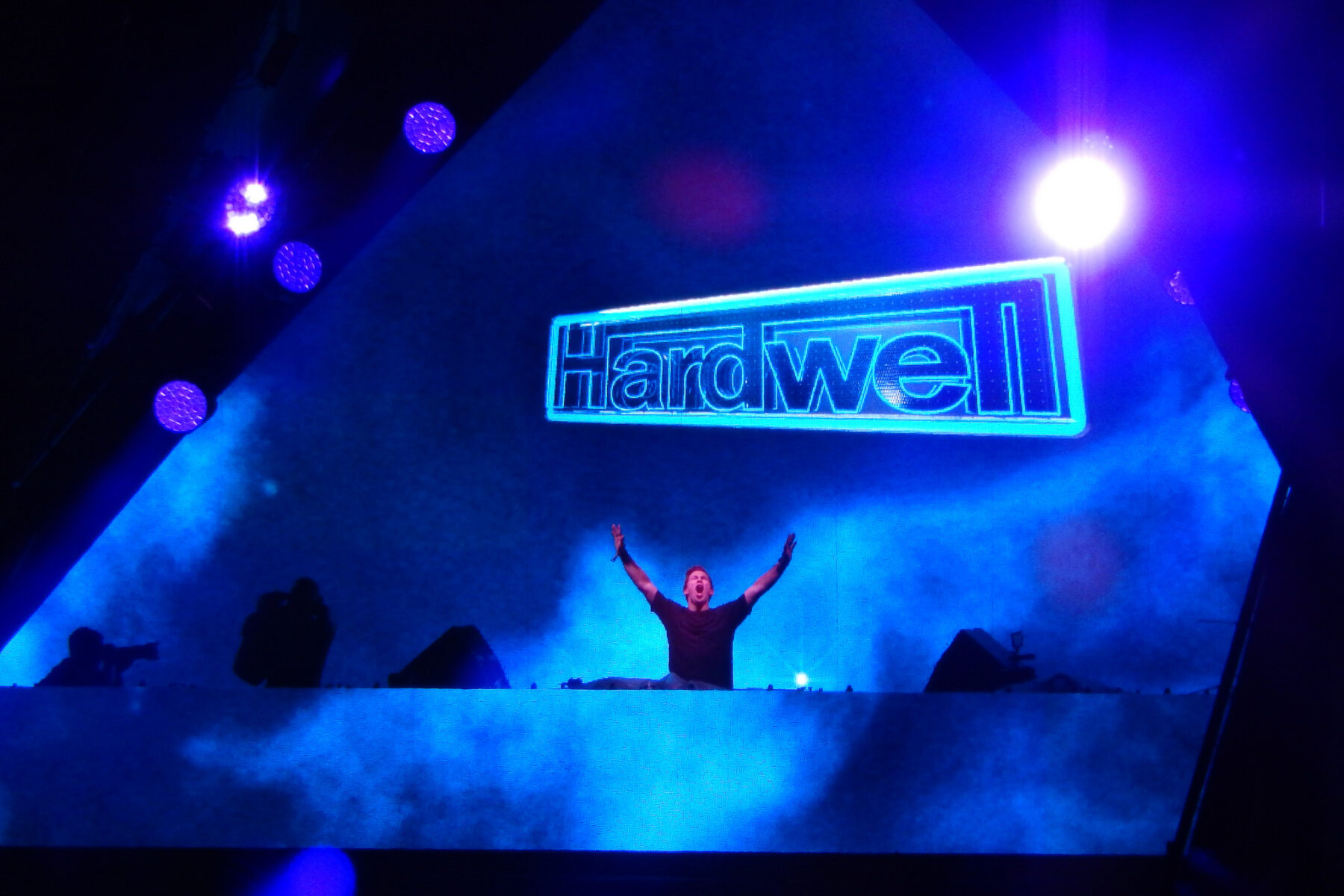 Hardwell raises both arms behind the decks under a large lit Hardwell sign on a blue-lit stage.