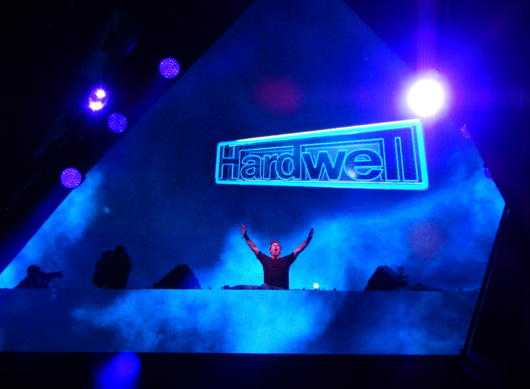 Hardwell raises both arms behind the decks under a large lit Hardwell sign on a blue-lit stage.