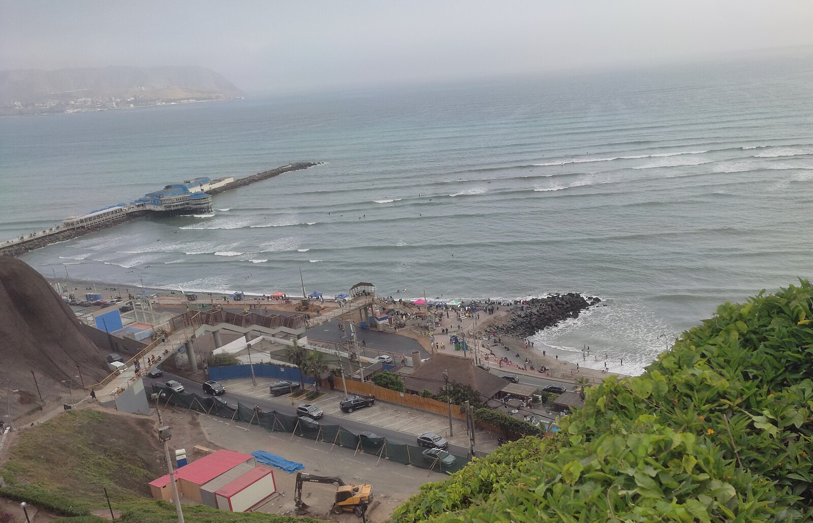View from the clifftop over a beach, parking and the coast road on Lima's Costa Verde, with surfers in the sea and a pier restaurant offshore.