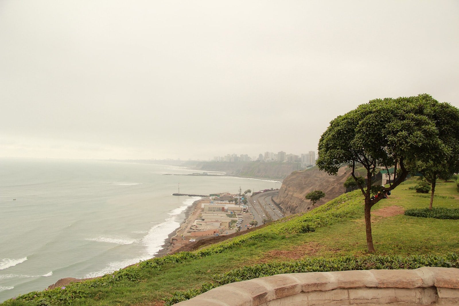 The cliffs of Lima's Costa Verde above the coast road and a strip of beach, with apartment towers on the clifftop in the distance under a grey sky.