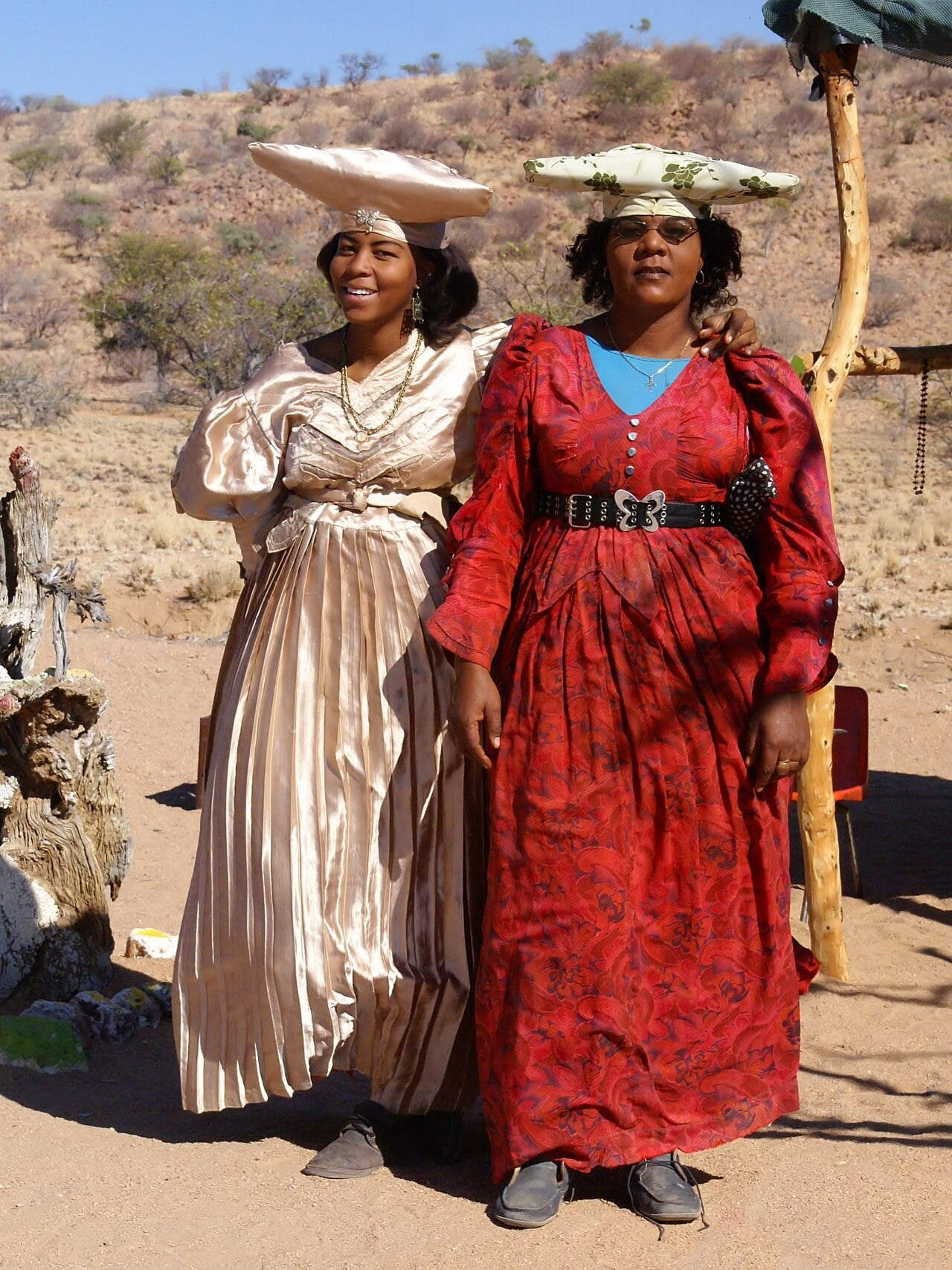 Two Herero women in long Victorian-style dresses and cloth headdresses standing on sandy ground in Namibia