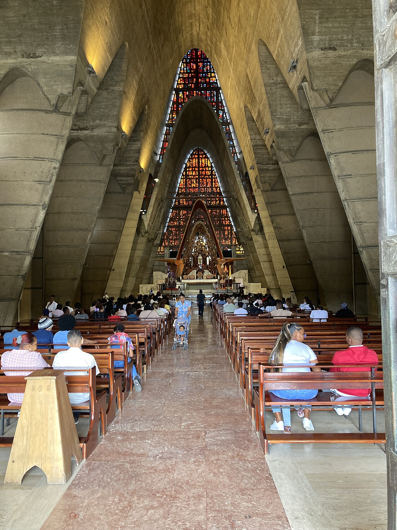Worshippers on wooden pews inside the Higüey basilica, facing stained-glass windows and the altar.