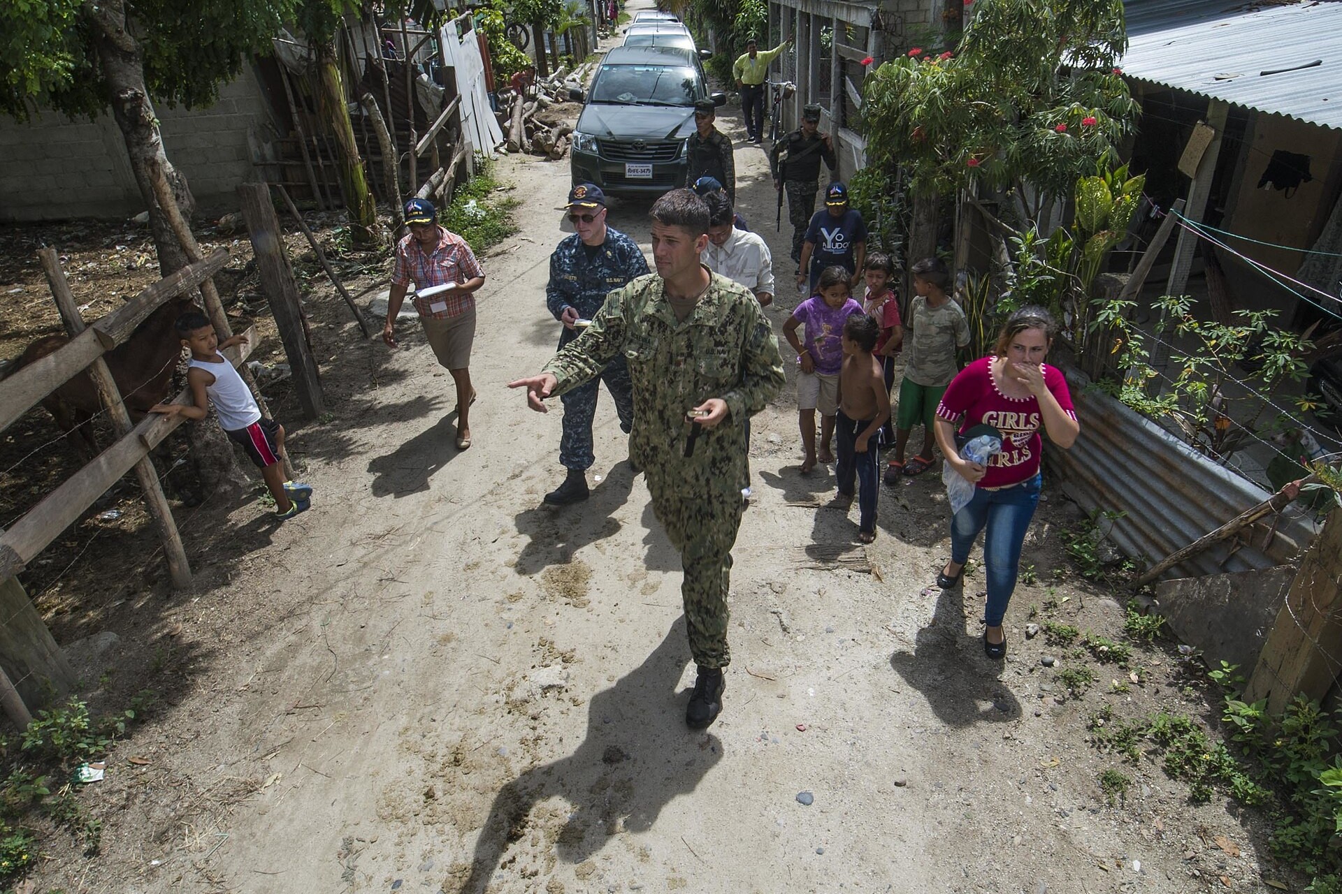 A US Navy officer in camouflage points down a dirt lane in Choloma, Honduras, followed by a Navy colleague, people with notebooks, Honduran soldiers and local children, during a mosquito-control house survey