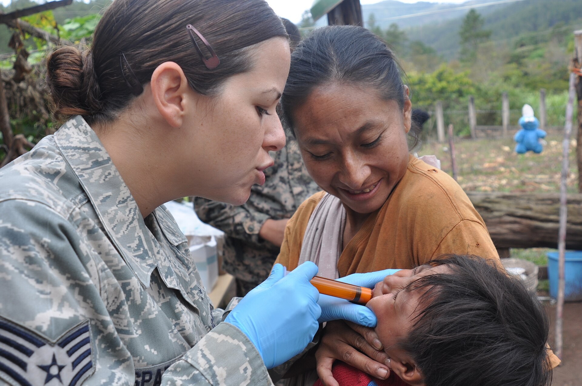 A US Air Force medic in uniform and blue gloves gives liquid medicine by syringe to a Honduran child held by a woman, with green hills in the background