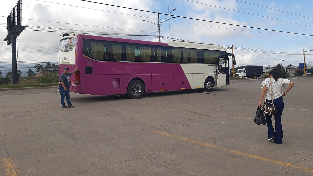 A purple and white long-distance coach manoeuvres on a concrete forecourt while a man guides it and a woman walks past with bags