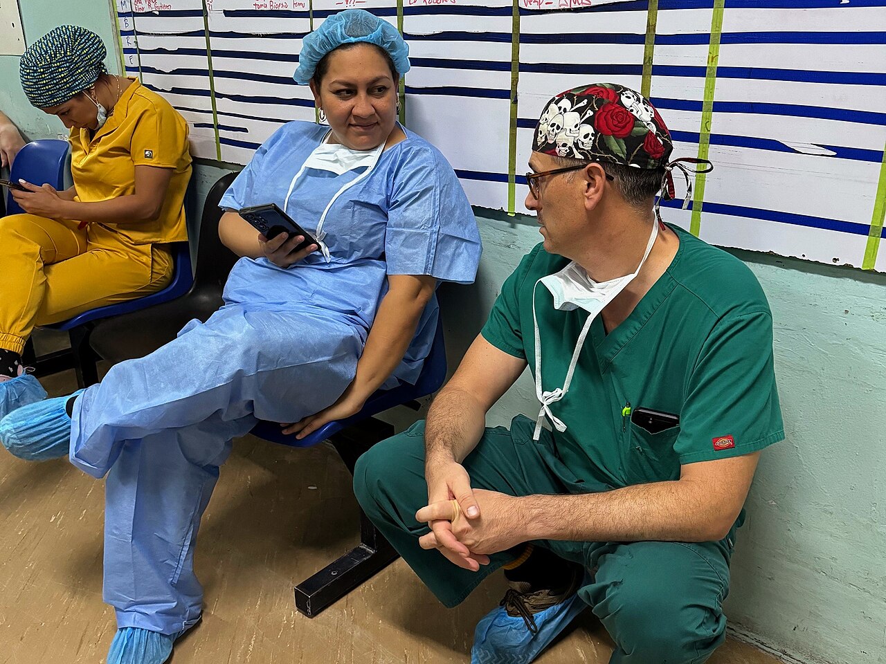 A Honduran surgeon in light blue scrubs and a hair cover talks with a visiting US Navy surgeon in green scrubs beside a surgery schedule board at Hospital Escuela