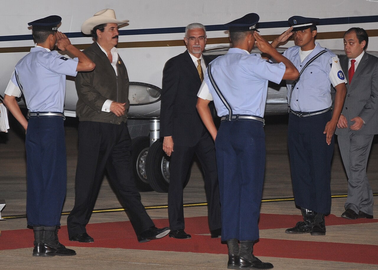 Manuel Zelaya in a white cowboy hat walks across a red carpet past saluting air force guards beside a small jet