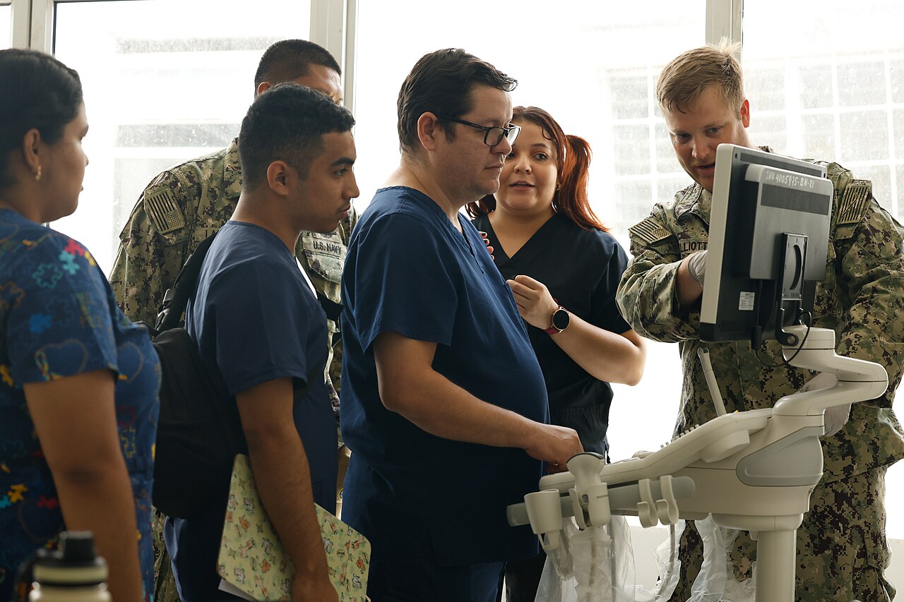 Medical staff in blue scrubs gather around an ultrasound machine as a US Navy technician explains its screen in a hospital ward