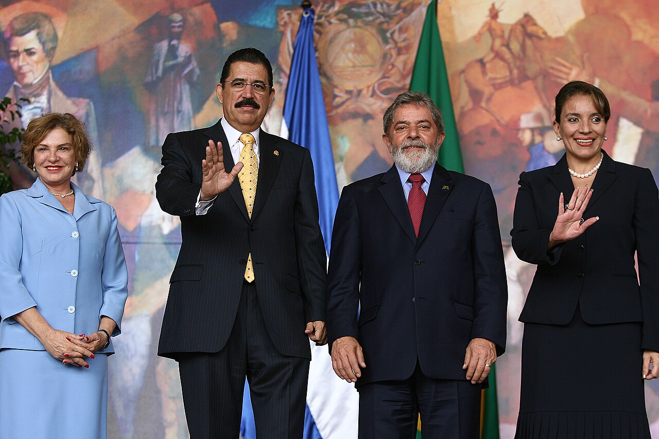 Manuel Zelaya in a dark suit and yellow tie waving, beside Brazil's Lula and two women, in front of a large historical mural and national flags
