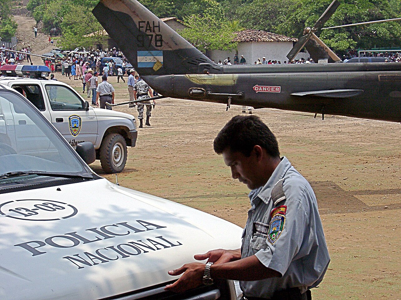 A Honduran police officer in a grey uniform leans on a white Policía Nacional pickup, with an air force helicopter and a crowd behind him