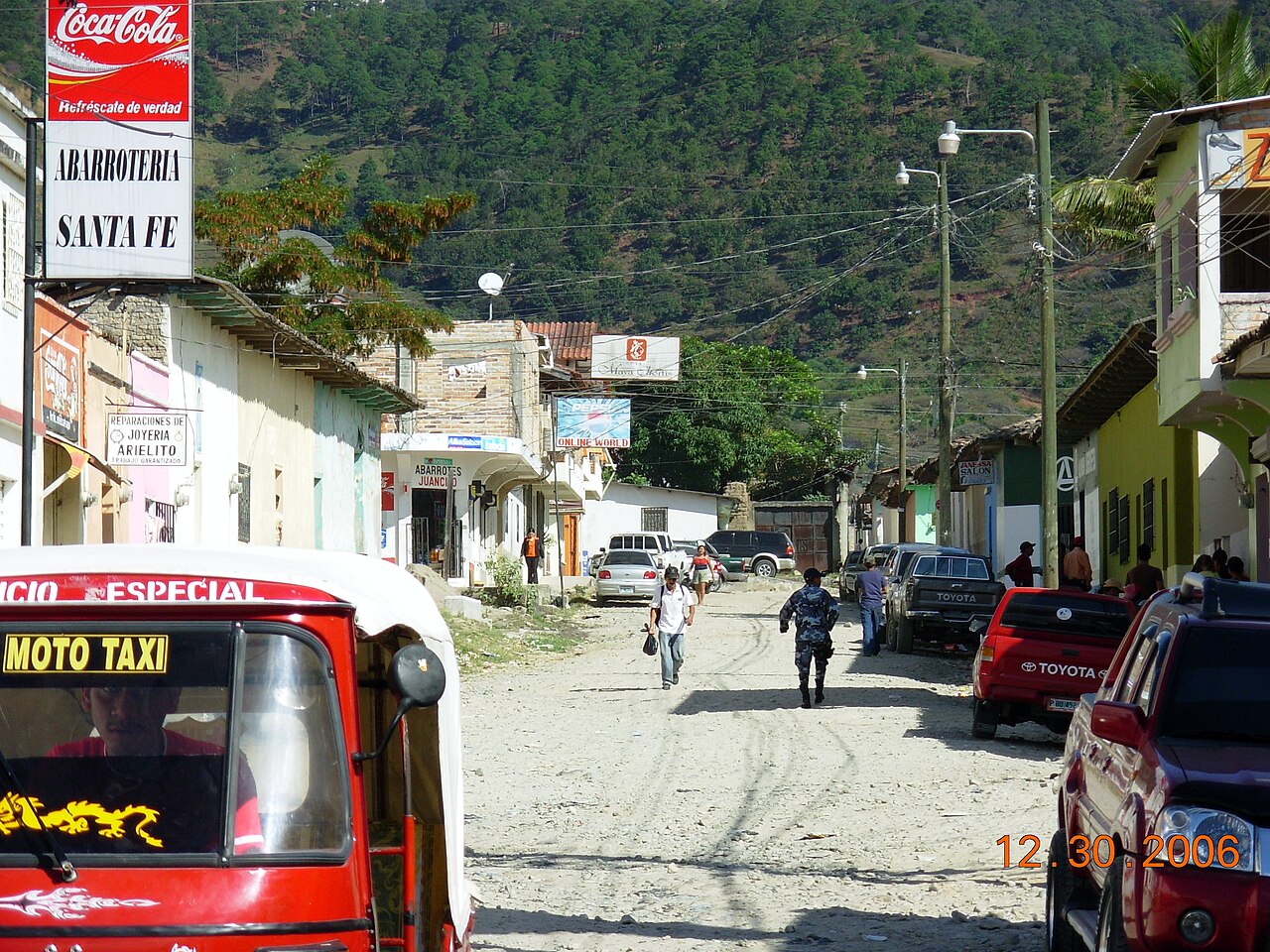 Unpaved street in Nueva Ocotepeque, Honduras, with a red moto-taxi, parked pickups, shops and pine-covered hills behind