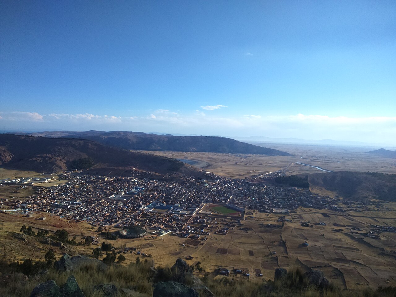 View from a hillside over the town of Huancané on the high plain of Peru's Puno region, with hills and brown farm fields around it
