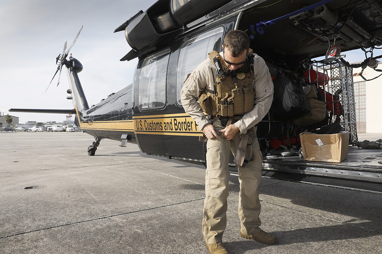 A US Customs and Border Protection aircrew member in tactical vest adjusts his gear beside a black Black Hawk helicopter on an airfield apron