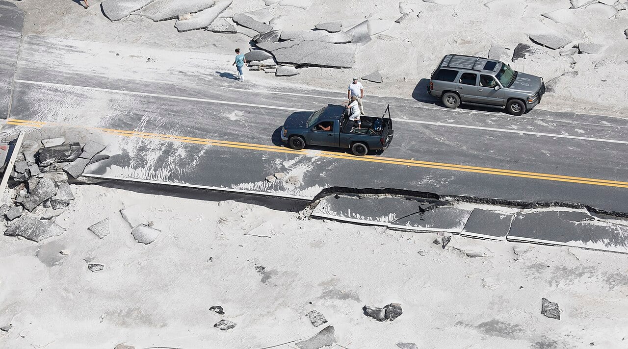 Aerial view of a coastal road on the Florida Panhandle covered in sand and broken asphalt, with two vehicles and several people standing on it after a hurricane
