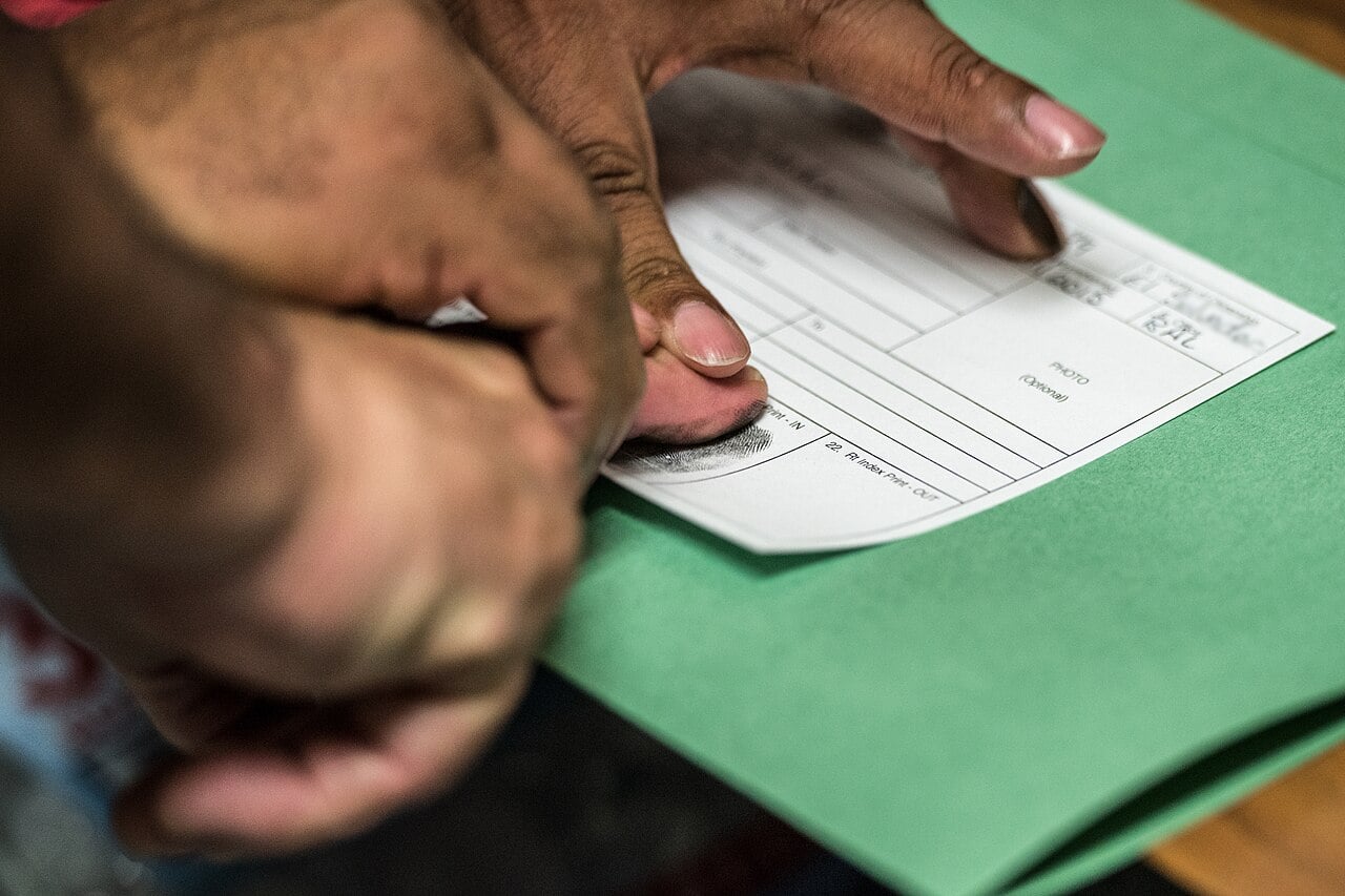 An immigration officer presses a suspect's fingers onto a fingerprint card on a desk