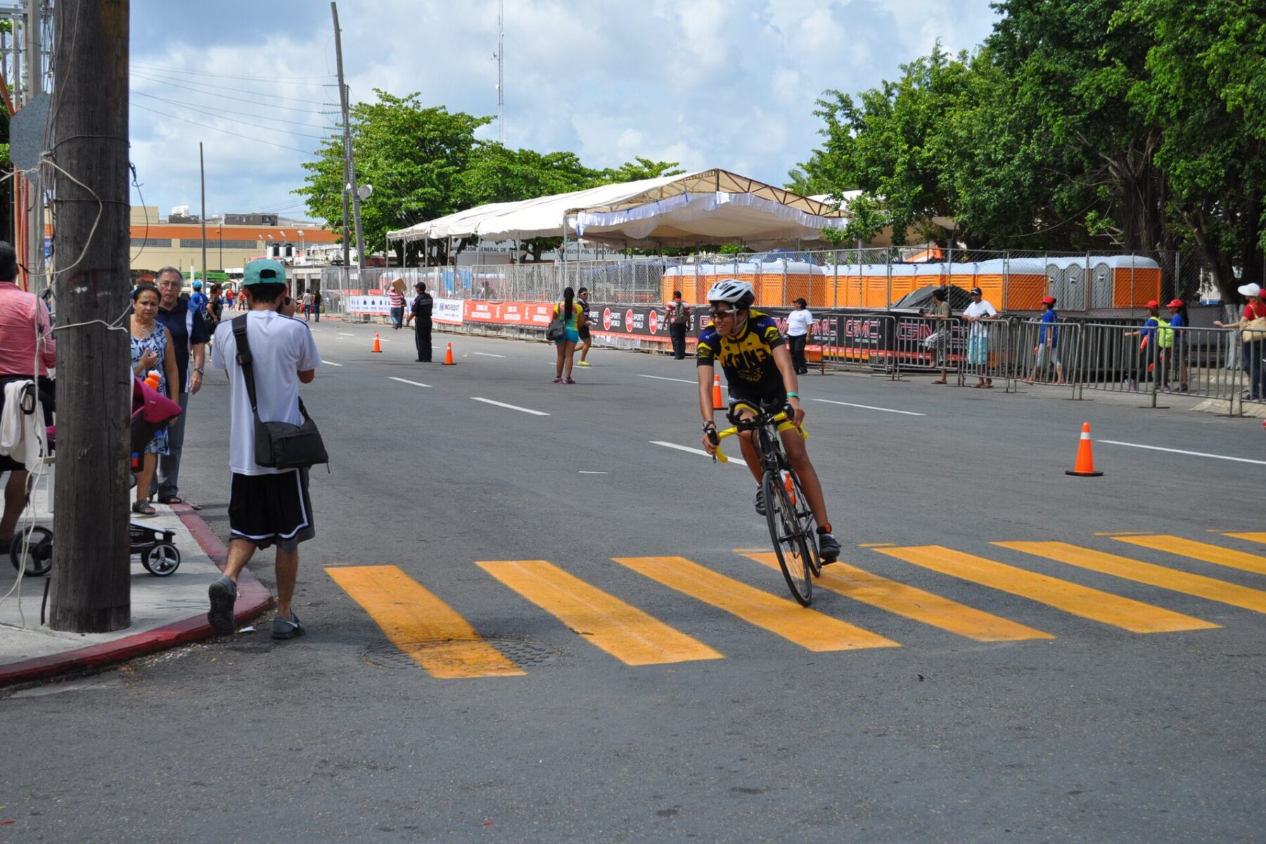 A triathlete in a yellow and black kit rides past spectators and barriers on a closed street in Cozumel