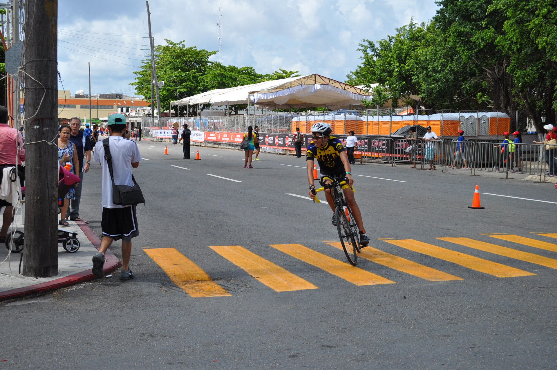 A triathlete in a yellow and black kit rides past spectators and barriers on a closed street in Cozumel