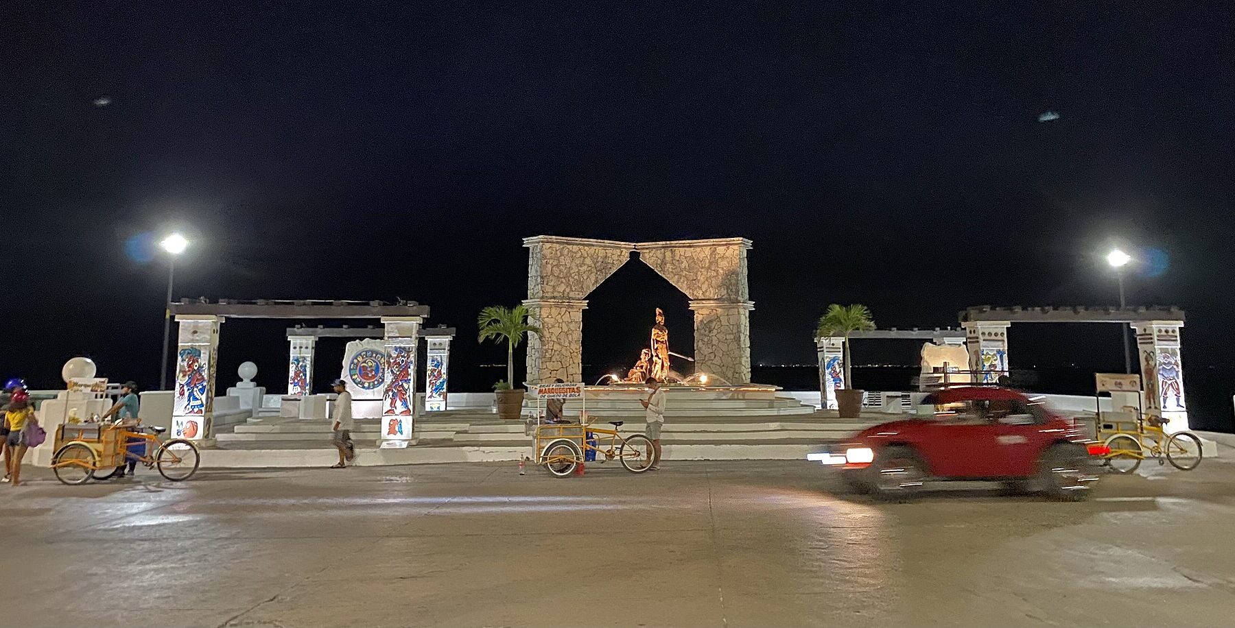 The Malecón waterfront in San Miguel de Cozumel at night, with a stone arch monument, bicycle carts and a red Volkswagen Beetle