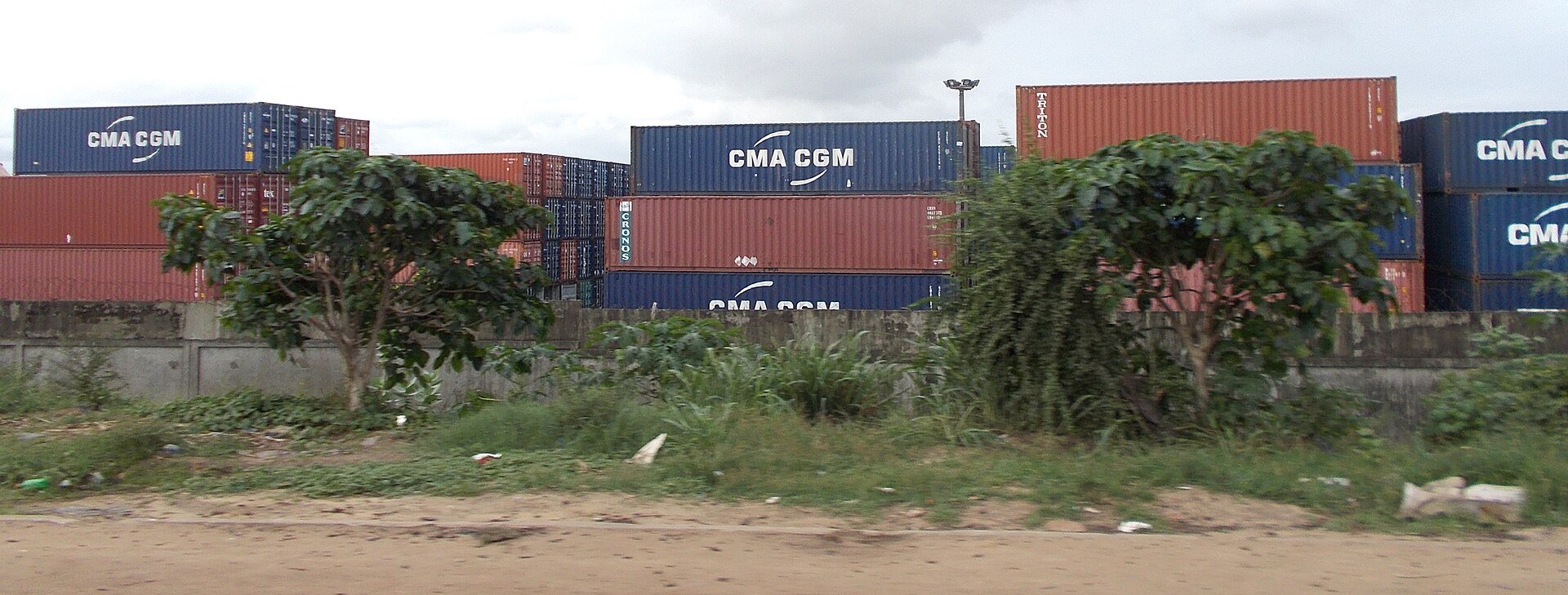 Stacks of blue and red shipping containers behind a wall and trees near the port of Abidjan, Ivory Coast