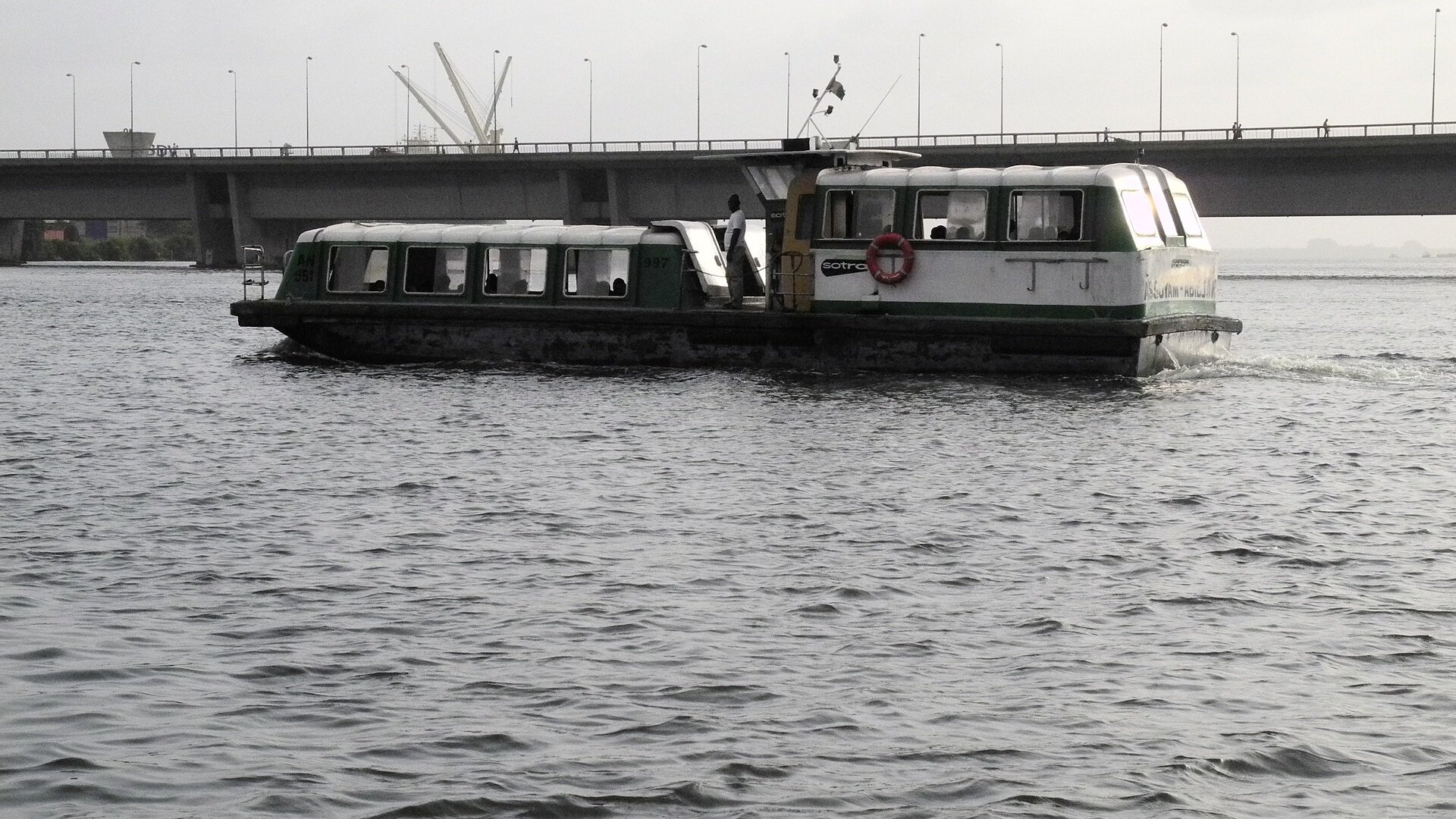 A green and white SOTRA water bus crossing the Ébrié Lagoon in Abidjan beneath a road bridge, with port cranes in the distance