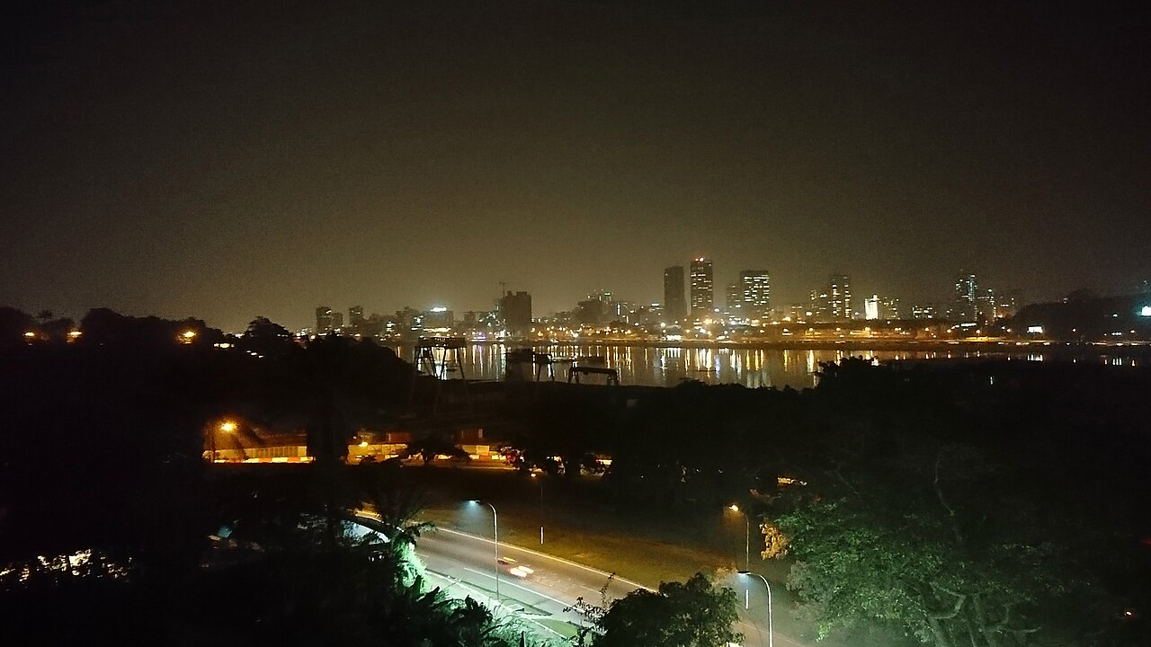 Lit towers of Abidjan's Plateau district reflected in the lagoon at night, with a road in the foreground