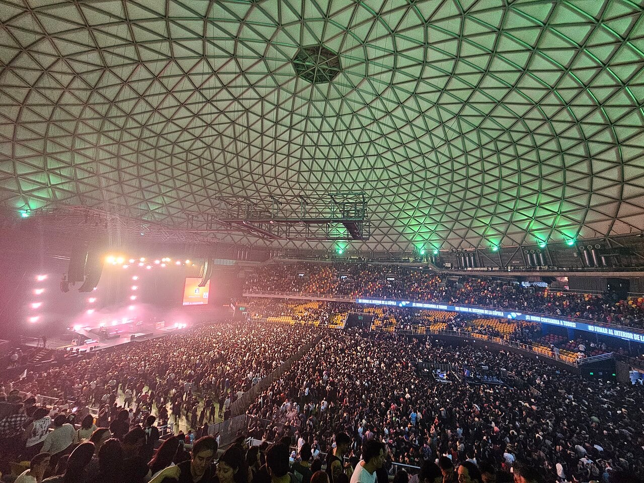 Packed concert crowd under the geodesic dome of the Movistar Arena in Santiago, Chile, with a lit stage at left.