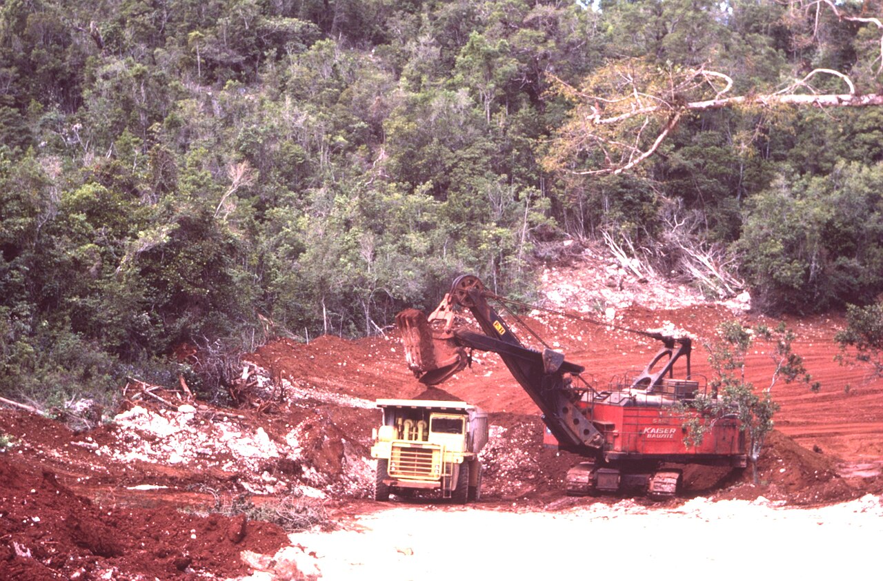 A red Kaiser Bauxite power shovel loading a dump truck at an open bauxite pit cut into forest in Jamaica