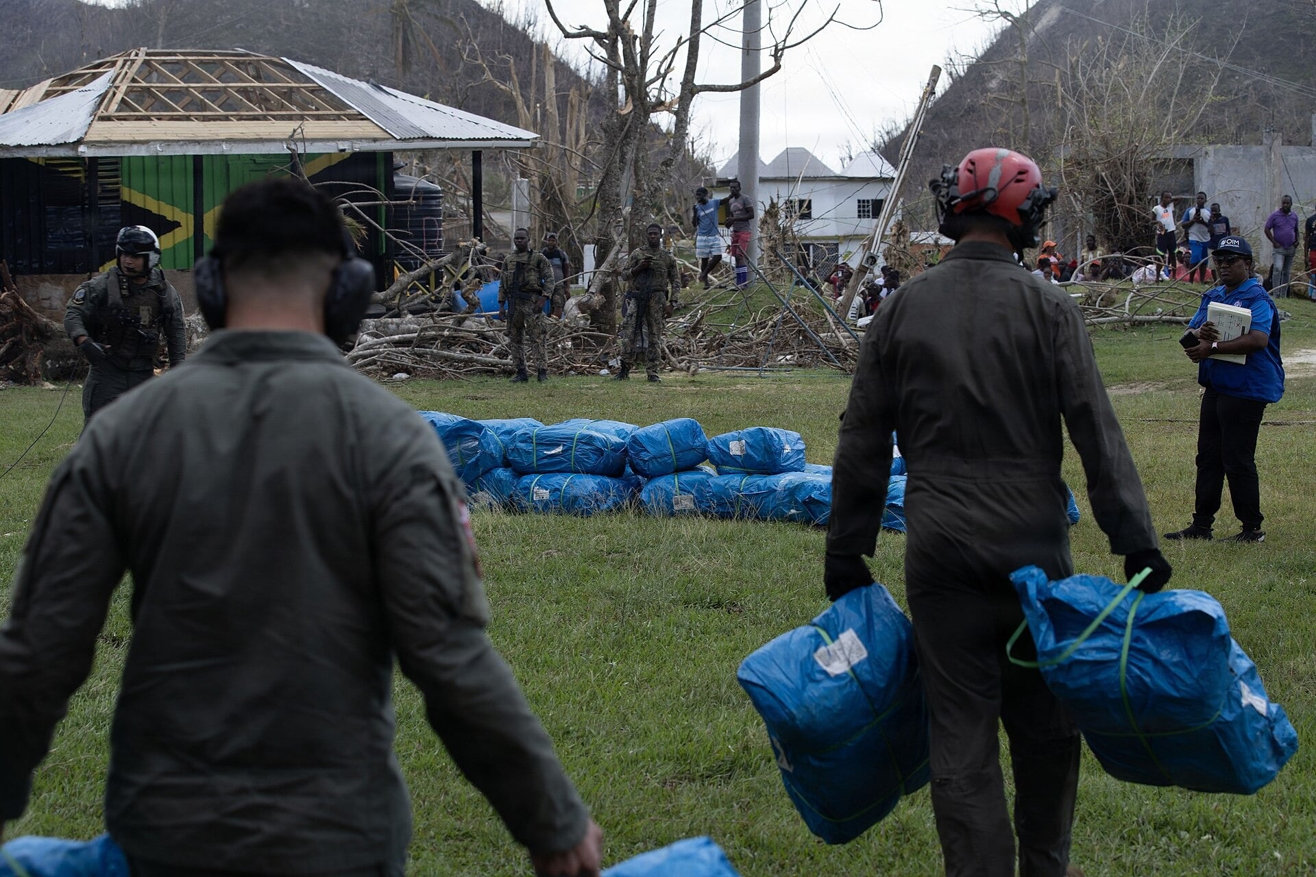 US Marines and aircrew carry blue tarpaulin bundles across a field in Beeston Spring, Jamaica, watched by Jamaican soldiers, beside a building whose roof frame is stripped bare
