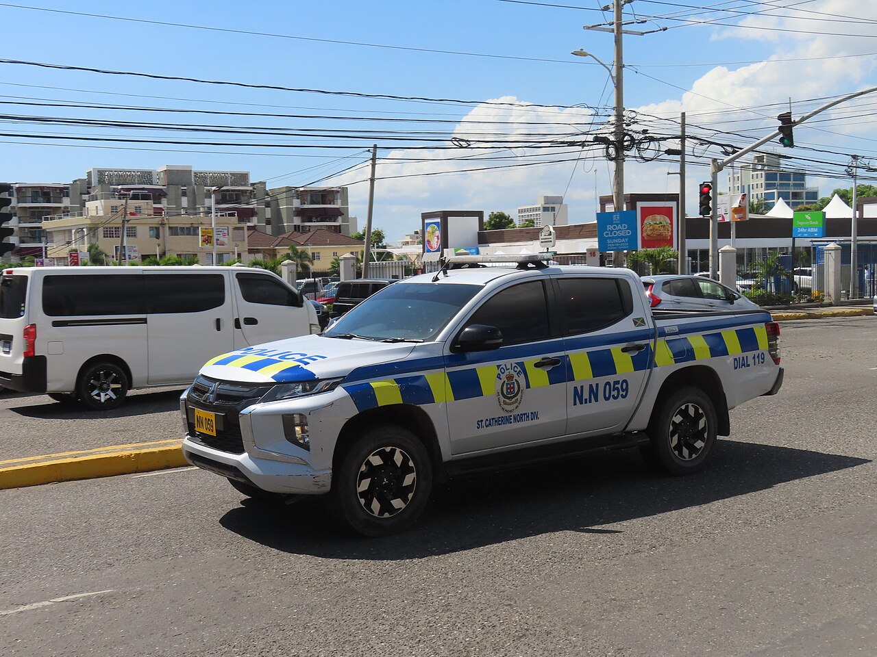 White Jamaica Constabulary Force pick-up truck with blue and yellow markings driving through a Kingston junction.