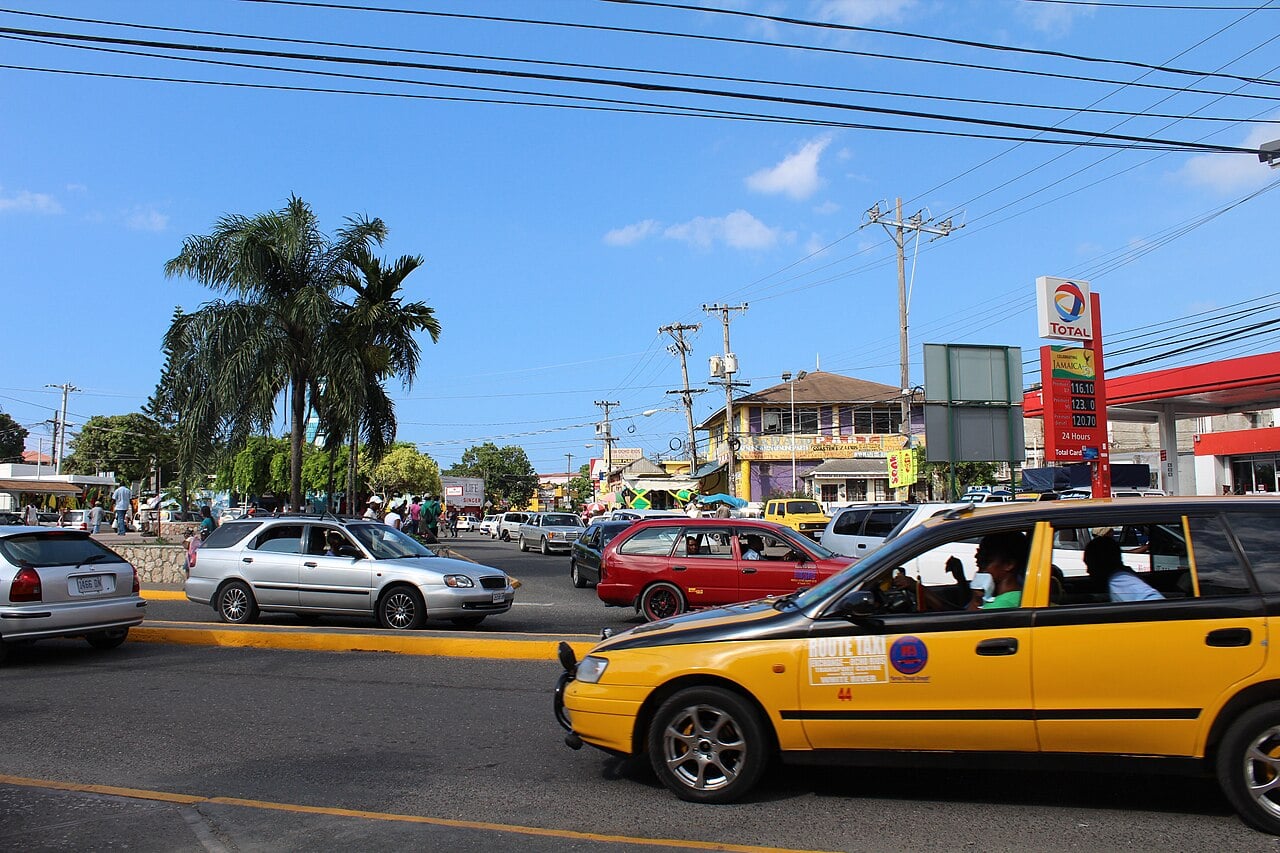 Taxis and cars at a busy junction in Kingston, Jamaica, with palm trees, a fuel station and shops under a blue sky