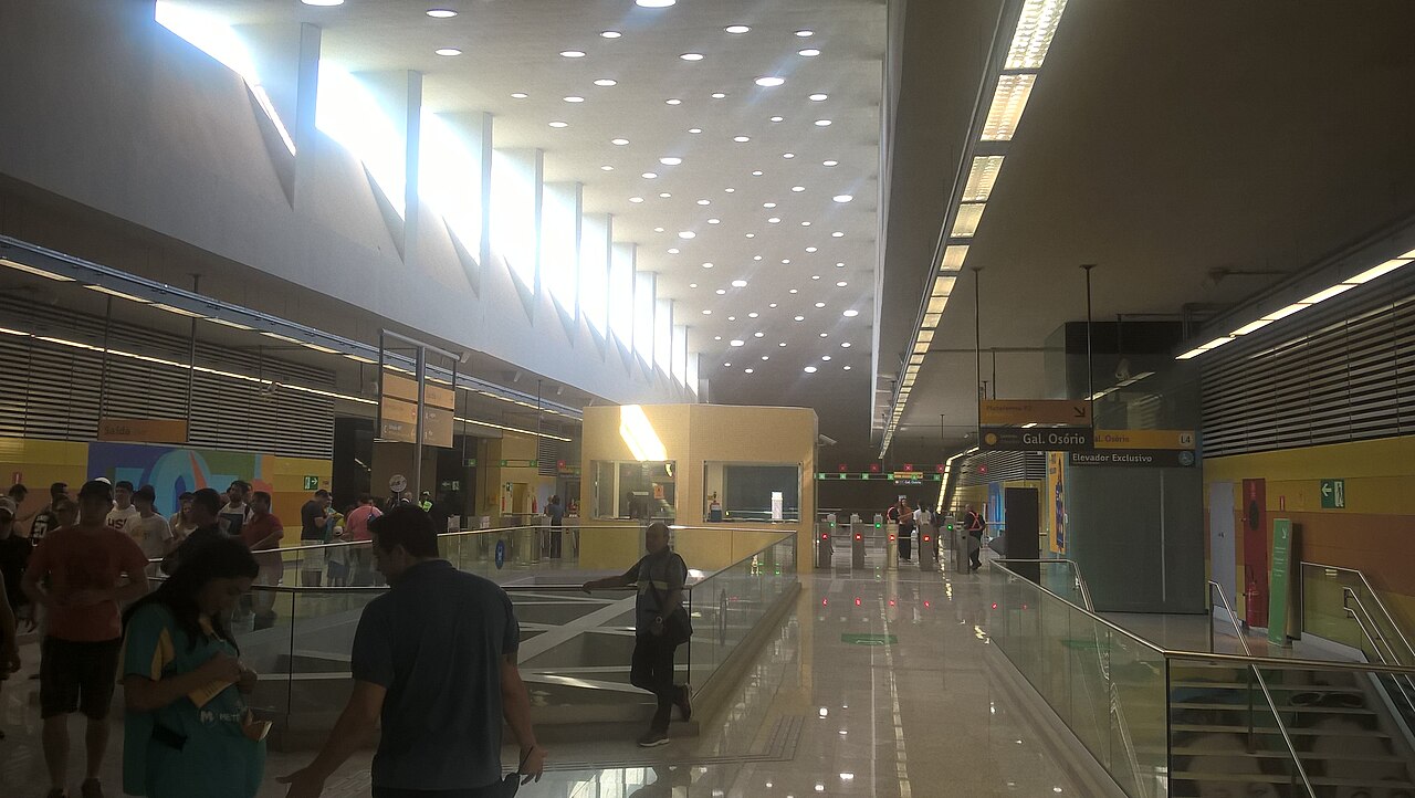 The main hall of Jardim Oceânico metro station in Barra da Tijuca, with ticket gates, yellow walls and a tall ceiling lit by skylights, as passengers walk through.