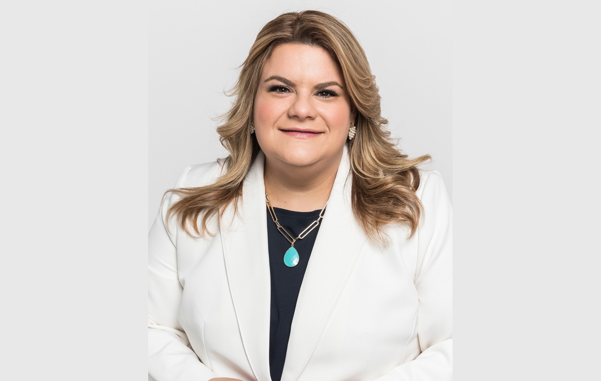 Puerto Rico Governor Jenniffer González-Colón smiling in a white blazer against a light grey studio background