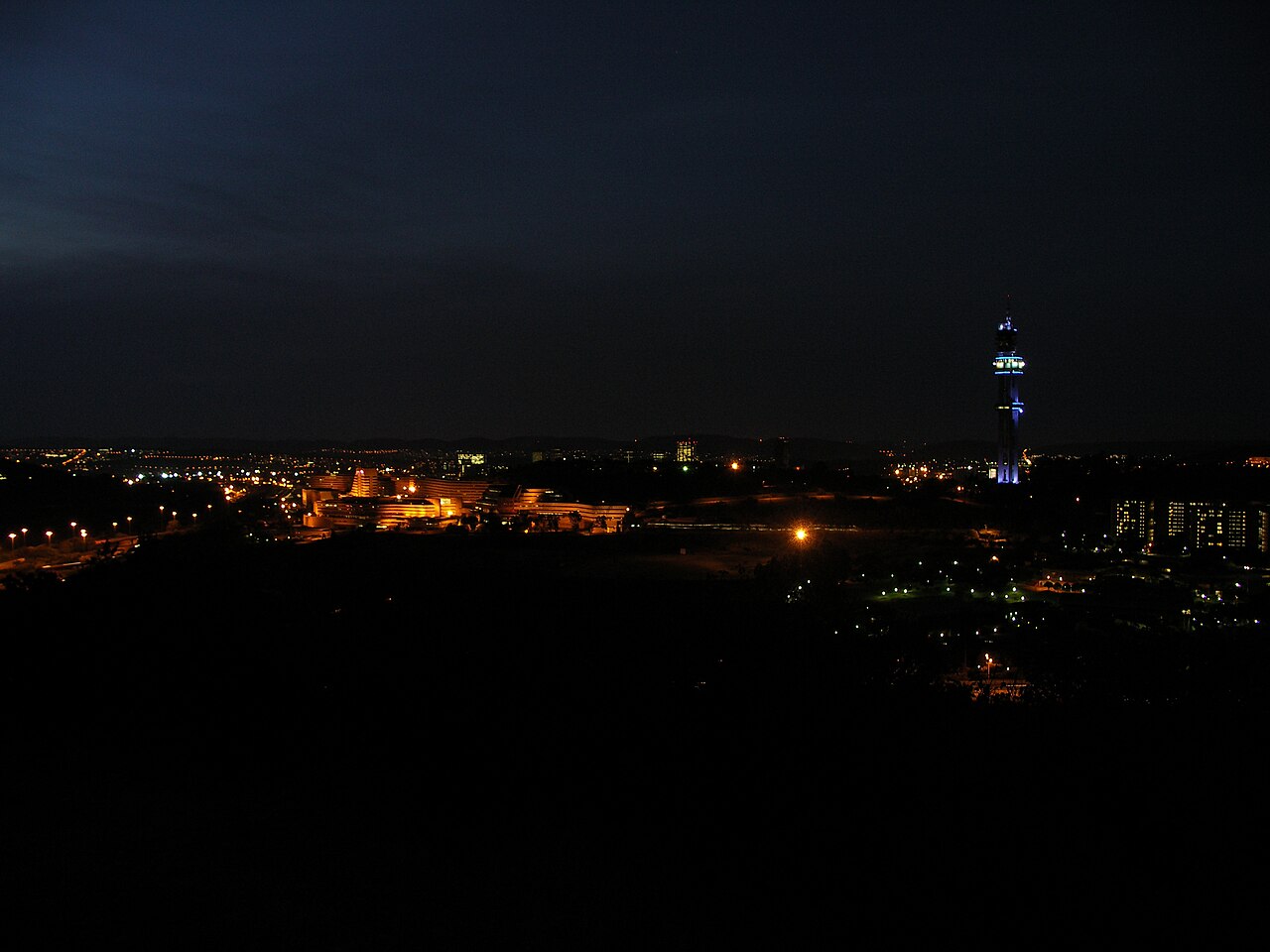 Pretoria at night seen from a hill, with city lights, a lit building complex and a tall telecommunications tower lit in blue against a dark sky.