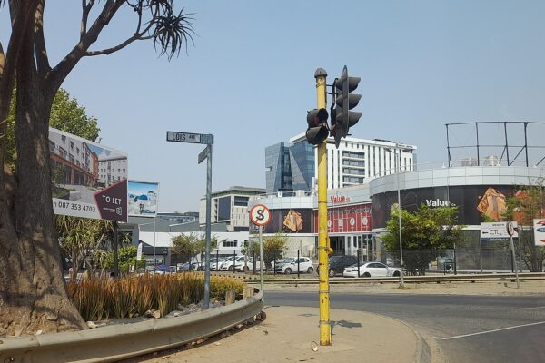 Lois Avenue in Menlyn, Pretoria, with traffic lights, a street sign, office blocks and a round shopping building under a clear sky.