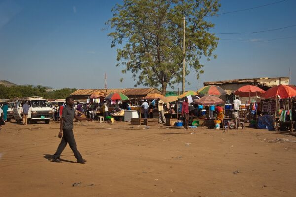 Market stalls under colourful umbrellas on a dusty square in Juba, South Sudan, with minibuses parked on the left