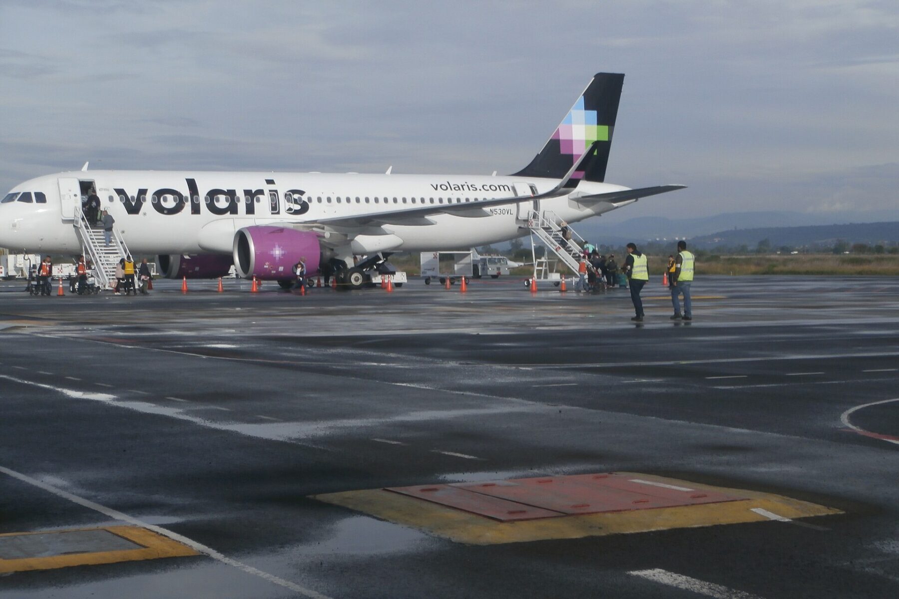 Passengers board a white Volaris jet with a purple engine by stairs on a wet apron under grey sky