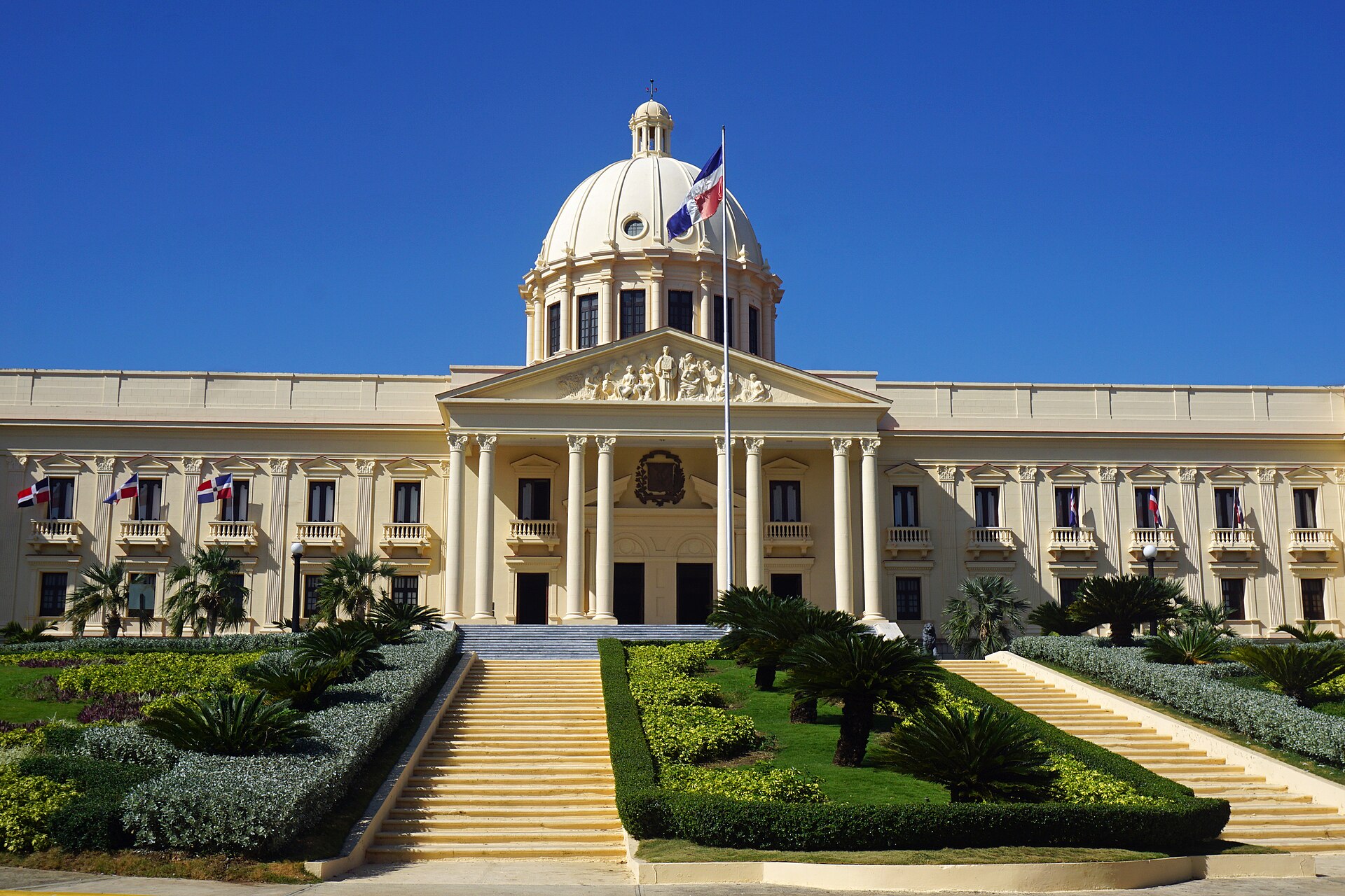A cream neoclassical palace with a white dome, columned portico and Dominican flags, with stairs and trimmed hedges in front under a deep blue sky