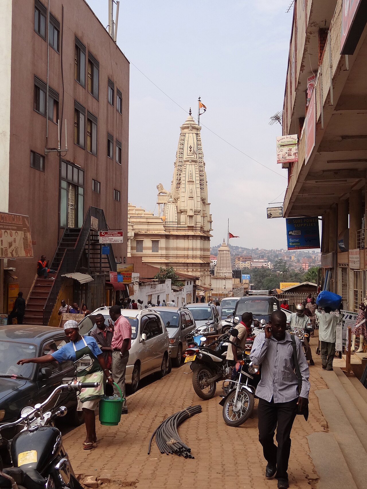 A busy downtown street in Kampala, Uganda, with parked cars, motorbike taxis, pedestrians and a white temple tower in the background
